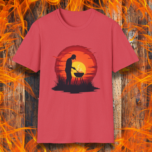 Heather Red  T-shirt featuring a vibrant design of a silhouetted man grilling in front of a large, glowing sunset. The sunset has warm hues of red, orange, and yellow, creating a striking contrast against the dark background. The scene is framed with grass and subtle clouds, giving it an outdoorsy, serene BBQ vibe.