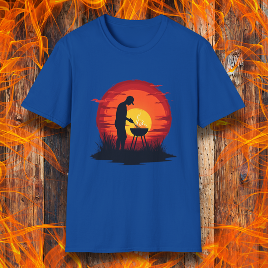 Royal Blue T-shirt featuring a vibrant design of a silhouetted man grilling in front of a large, glowing sunset. The sunset has warm hues of red, orange, and yellow, creating a striking contrast against the dark background. The scene is framed with grass and subtle clouds, giving it an outdoorsy, serene BBQ vibe.