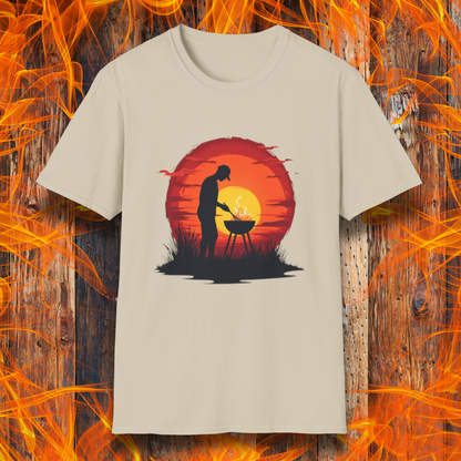 Sand  T-shirt featuring a vibrant design of a silhouetted man grilling in front of a large, glowing sunset. The sunset has warm hues of red, orange, and yellow, creating a striking contrast against the dark background. The scene is framed with grass and subtle clouds, giving it an outdoorsy, serene BBQ vibe.