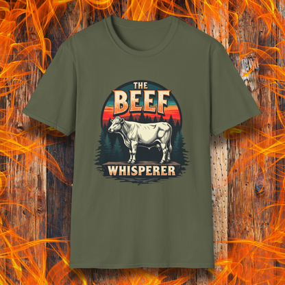 Military Green T-shirt featuring a bold design with the words "The Beef Whisperer" in a vintage western-style font. Above the text, a detailed illustration of a cow stands on a patch of ground, framed by a scenic forest and a sunset with deep orange and red hues. The design has a rustic, outdoorsy feel, appealing to BBQ lovers and beef enthusiasts.