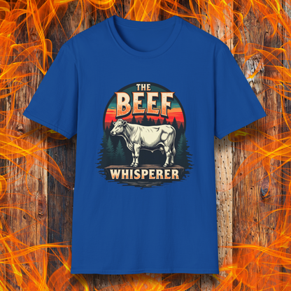 Royal Blue  T-shirt featuring a bold design with the words "The Beef Whisperer" in a vintage western-style font. Above the text, a detailed illustration of a cow stands on a patch of ground, framed by a scenic forest and a sunset with deep orange and red hues. The design has a rustic, outdoorsy feel, appealing to BBQ lovers and beef enthusiasts.