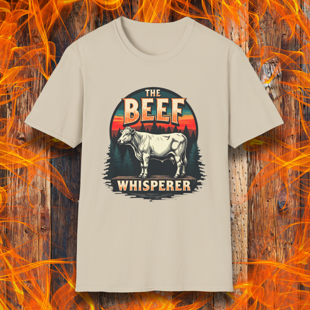 Sand  T-shirt featuring a bold design with the words "The Beef Whisperer" in a vintage western-style font. Above the text, a detailed illustration of a cow stands on a patch of ground, framed by a scenic forest and a sunset with deep orange and red hues. The design has a rustic, outdoorsy feel, appealing to BBQ lovers and beef enthusiasts.
