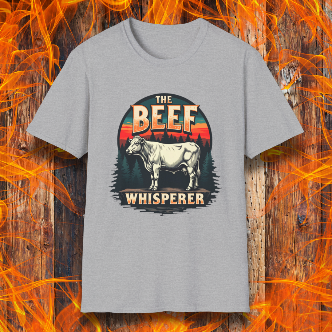 Sport Grey  T-shirt featuring a bold design with the words "The Beef Whisperer" in a vintage western-style font. Above the text, a detailed illustration of a cow stands on a patch of ground, framed by a scenic forest and a sunset with deep orange and red hues. The design has a rustic, outdoorsy feel, appealing to BBQ lovers and beef enthusiasts.