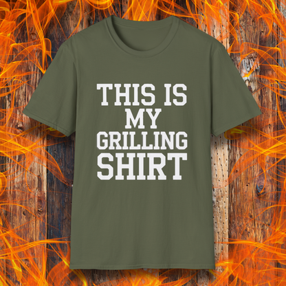 Military Green T-shirt with bold white text that reads "THIS IS MY GRILLING SHIRT." The shirt is displayed against a fiery wooden background, emphasizing its BBQ and grilling theme.