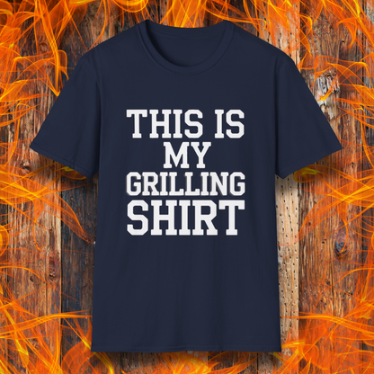 Navy T-shirt with bold white text that reads "THIS IS MY GRILLING SHIRT." The shirt is displayed against a fiery wooden background, emphasizing its BBQ and grilling theme.