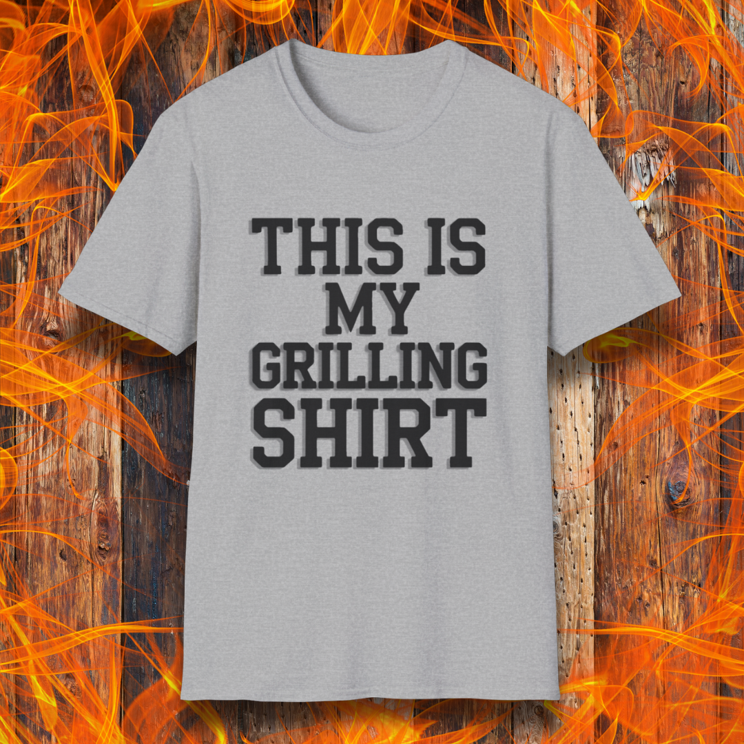 Sport Grey  T-shirt with bold black text that reads "THIS IS MY GRILLING SHIRT." The shirt is displayed against a fiery wooden background, emphasizing its BBQ and grilling theme.