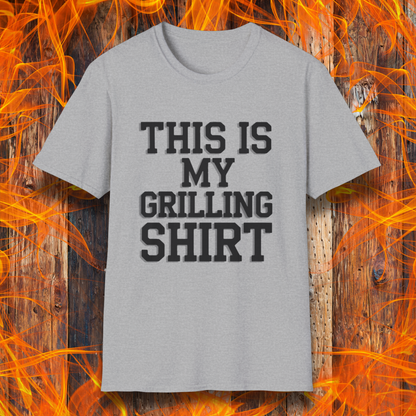 Sport Grey  T-shirt with bold black text that reads "THIS IS MY GRILLING SHIRT." The shirt is displayed against a fiery wooden background, emphasizing its BBQ and grilling theme.